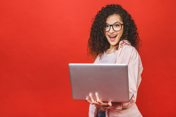 Portrait of an excited curly young girl holding laptop computer isolated over red background.