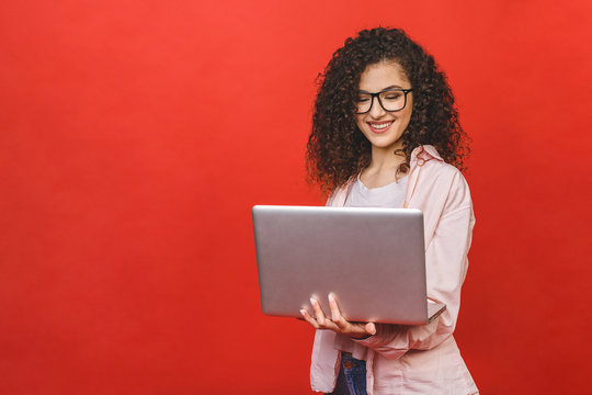 Portrait of an excited curly young girl holding laptop computer isolated over red background.