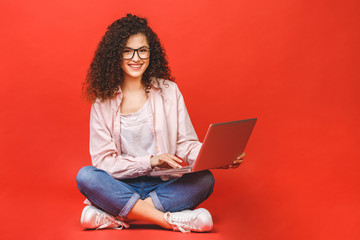 Happy young curly beautiful woman sitting on the floor with crossed legs and using laptop on red...