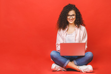 Happy young curly beautiful woman sitting on the floor with crossed legs and using laptop on red...