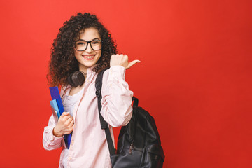 Young curly student woman wearing backpack glasses holding books and tablet over isolated red background. Pointing finger. - obrazy, fototapety, plakaty