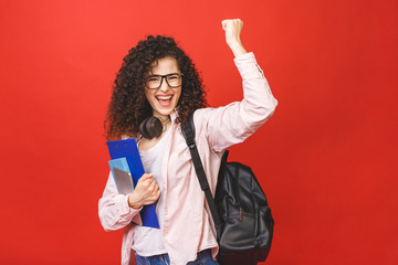 Happy winner celebrating victory. Young curly student woman wearing backpack glasses holding books and tablet over isolated red background. Pointing finger. - obrazy, fototapety, plakaty