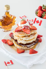 A vertical close up view of a plate of pancakes with strawberries with maple syrup in behind. 