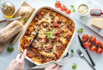 Top down view of a freshly baked lasagna being cut into and surrounded by Italian ingredients.