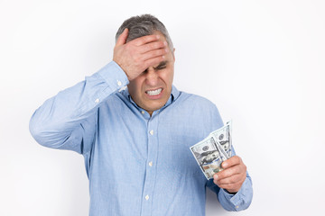 adult upset man with grey hair wearing blue shirt holds cash in his hand looks regretful standing...