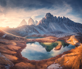 Aerial view of beautiful rocks, mountain lake, reflection in water and houses on the hill at sunset. Autumn landscape with mountains, blue sky and sunlight. Dolomites, Italy. Top view of Italian alps