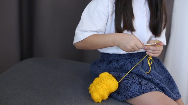 Little girl sitting on couch and knitting wool scarf. Young girl using crochet for knitting wool clothes in home room. Girl making handmade clothes.