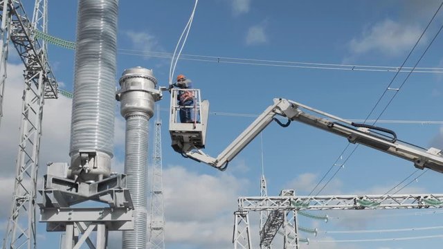 worker installs modern equipment standing on truck crane mounting basket at high voltage power transmission tower under blue sky