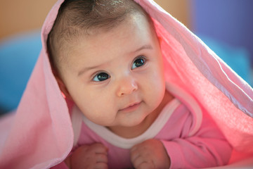 Cute three months old Baby girl infant on a bed on her belly with head up looking into camera with her big eyes. Natural bedroom light.