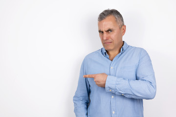 mature handsome confused man with grey hair wearing blue shirt standing on isolated white background points with his fingers looking bewildered, body language concept