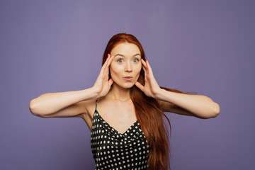 Surprised happy young female stands on purple background, glad to see big discounts on clothes, expresses shock, keeps hands near head, jaw dropped, going to spend much money today