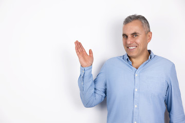 Fototapeta premium mature smiling man with grey hair wearing blue shirt standing on isolated white background holds his hand up and to the side, body language concept