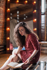 Portrait of attractive student girl with curly hair, in short red dress and round glasses. Young woman sitting on glass coffee table in loft apartment and reading book in her hands.