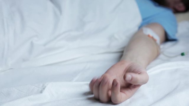 Terminally ill Caucasian male patients hand convulsing, dropper in arm, person suffering pain on a hospital bed