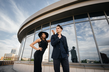 A beautiful, stylish pair of young people in black clothes and glasses stand against the background of an office building in the sunset. Fashion and style