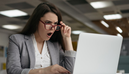 Portrait of shocked business woman in stylish eyeglasses using laptop computer , looking at digital...