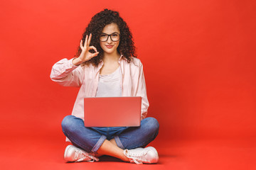 Happy young curly beautiful woman sitting on the floor with crossed legs and using laptop on red background. Ok sign. - obrazy, fototapety, plakaty