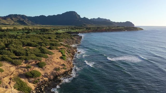 Low elevation aerial video of the southeast coast of Kauai with Lithified cliffs covered with ironwood pine trees, green golf courses and mountain range. Poipu, Koloa, Kauai