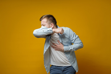 Man in a medical mask coughs in his elbow and holds hand on chest, experiencing pain. Concept of...
