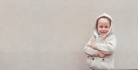 Happy joyful little boy 4-5 years old, standing in beige jumper with hood, background wall, cheerful on positive. Free space for copy text, banner for tagline. Emotions relaxation fun joy celebration.