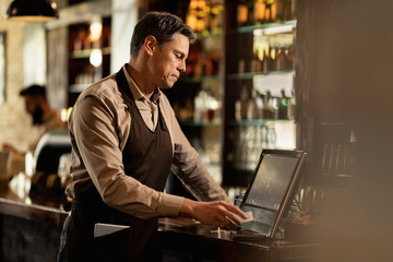 Male waiter going through orders while working at cash register in a bar. - obrazy, fototapety, plakaty