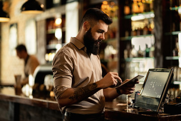 Young barista using touchpad while standing at cash register in a bar. - obrazy, fototapety, plakaty