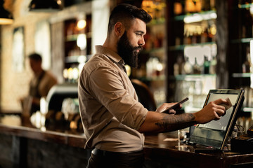Young bartender using cash register while working in a pub. - obrazy, fototapety, plakaty