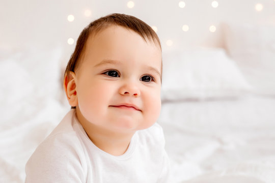 healthy baby boy 10 months old in white clothes smiling sitting on white bed linen in bed, space for text