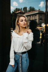 Beautiful stylish blonde woman in white shirt and jeans in city posing for camera. She is happy and enjoying walk in sunny day. She is wearing small white bag. Sarajevo, 2019.