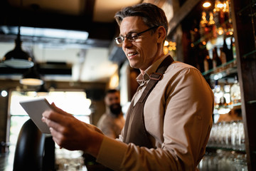 Happy mid adult waiter using digital tablet while working in a pub.