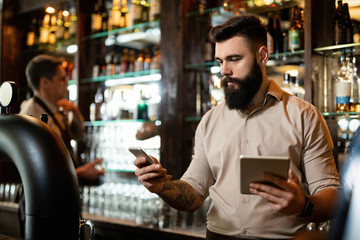 Fototapeta premium Young barista texting on the phone while using touchpad in a pub.