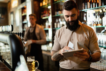 Happy bartender using digital tablet while working in a pub.