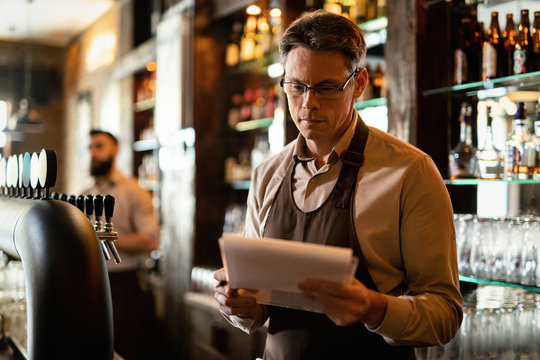 Smiling barista reading inventory list while working at bar counter in a bar.