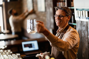 Happy bartender cleaning drinking glass while working at bar counter.
