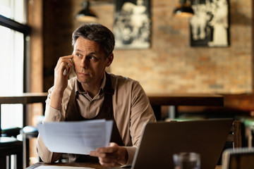 Mid adult waiter communicating on cell phone while analyzing paperwork in a pub.