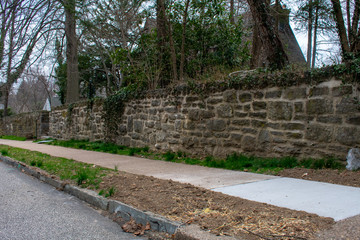 A Cobblestone Wall Next to a Paved Sidewalk and Road With Trees Behind It