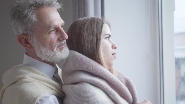 Close-up side view of young pretty Caucasian woman and senior man talking and smiling. Joyful married couple with age difference chatting indoors. Love, unity, leisure, lifestyle.
