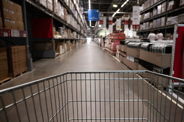 Fototapeta premium Empty shopping cart in a furniture store warehouse. Supermarket shopping basket aisle with box shelf abstract blur defocused background