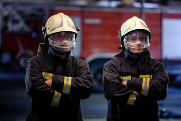 Fototapeta premium Two professional firemen posing. Firefighters wearing uniforms and protective helmets. Fire trucks in the background.