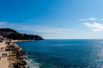 Panoramic wide angle shot of the Mediterranean Sea coastline leading to the lighthouse with the sunlight reflecting in the turquoise water and an airplane and seagulls flying above (Nice, France)
