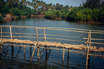 Old wooden bridge over a small river or pond, side view. Pedestrian bridge over the river on the background of green palm trees.