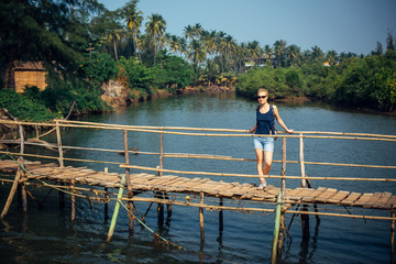 Fototapeta na wymiar A female tourist in denim shorts with short haircut on sunny tropical day. Young pretty girl stands on wooden bridge over small river against palm jungle and blue sky. Trips to picturesque places.