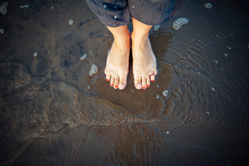 Woman's feet on sandy beach, standing in ocean water, copy space. Pedicured feet with red nail polish on toes in the sea. Bare feet standing on sand, top view. Hello summer, welcome vacation concept.