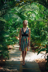 Slender beautiful woman in multicolored dress walking among tropical greenery on a hot sunny day. Attractive young girl enjoys privacy and relaxation in a Spa resort in Bali.