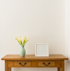White calla lilies in teal vase with blank square picture frame on oak side table against white wall