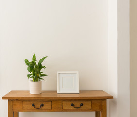 Green leafy houseplant in white pot with blank square picture frame on oak side table against white wall