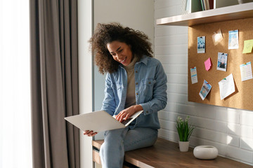 Young happy curly african girl woman lady student blogger freelancer sit table look at modern...