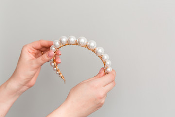 Close up of hands showing a trendy bridal gold diadem (hair hood, hair band, tiara) with big pearls. Wedding accessories. Copy space. Gray background. © Мария Крузман