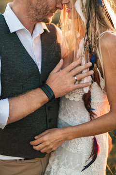 Cropped image of stylish rustic couple, boho woman in white dress and man in shirt, vest and pants posing in sunny field, hugging each other