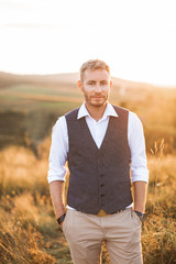 Portrait of handsome retro stylish man, posing on the background of summer field, sunset. Young bearded blond man wearing shirt, vest and pants, standing outdoors in the summer field